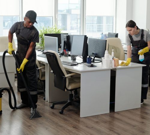 Young male and female workers of service company using detergent and vacuum cleaner for cleaning contemporary openspace office